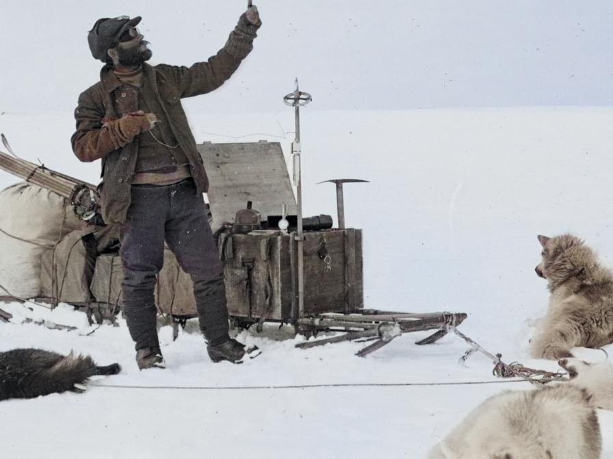 Mand, i gammeldags varmt tøj, foran en kasse på en slæde, med sne i bagrunden og to slædehunde der  hviler sig i sneen